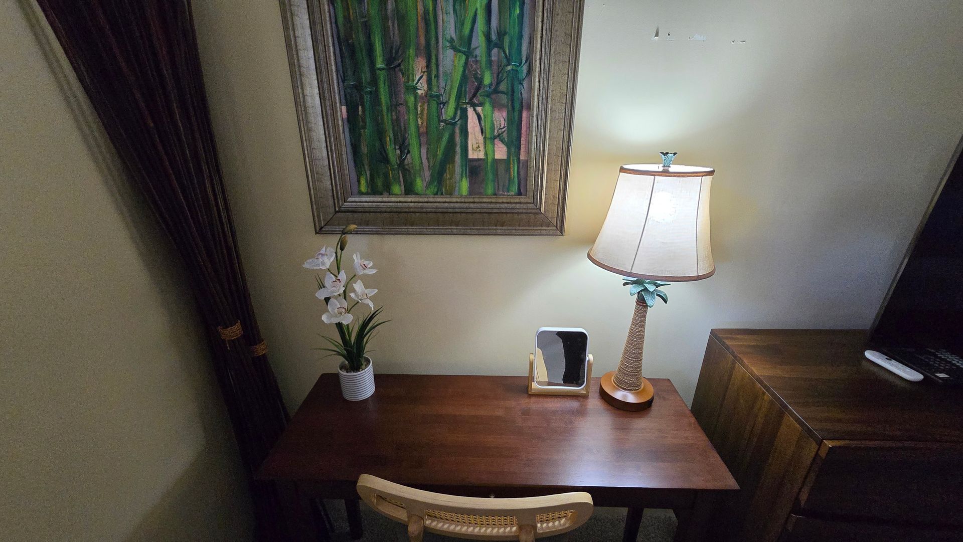 Desk with lamp, painting, flowers, and tablet. Wooden furniture in a room with a light beige wall.