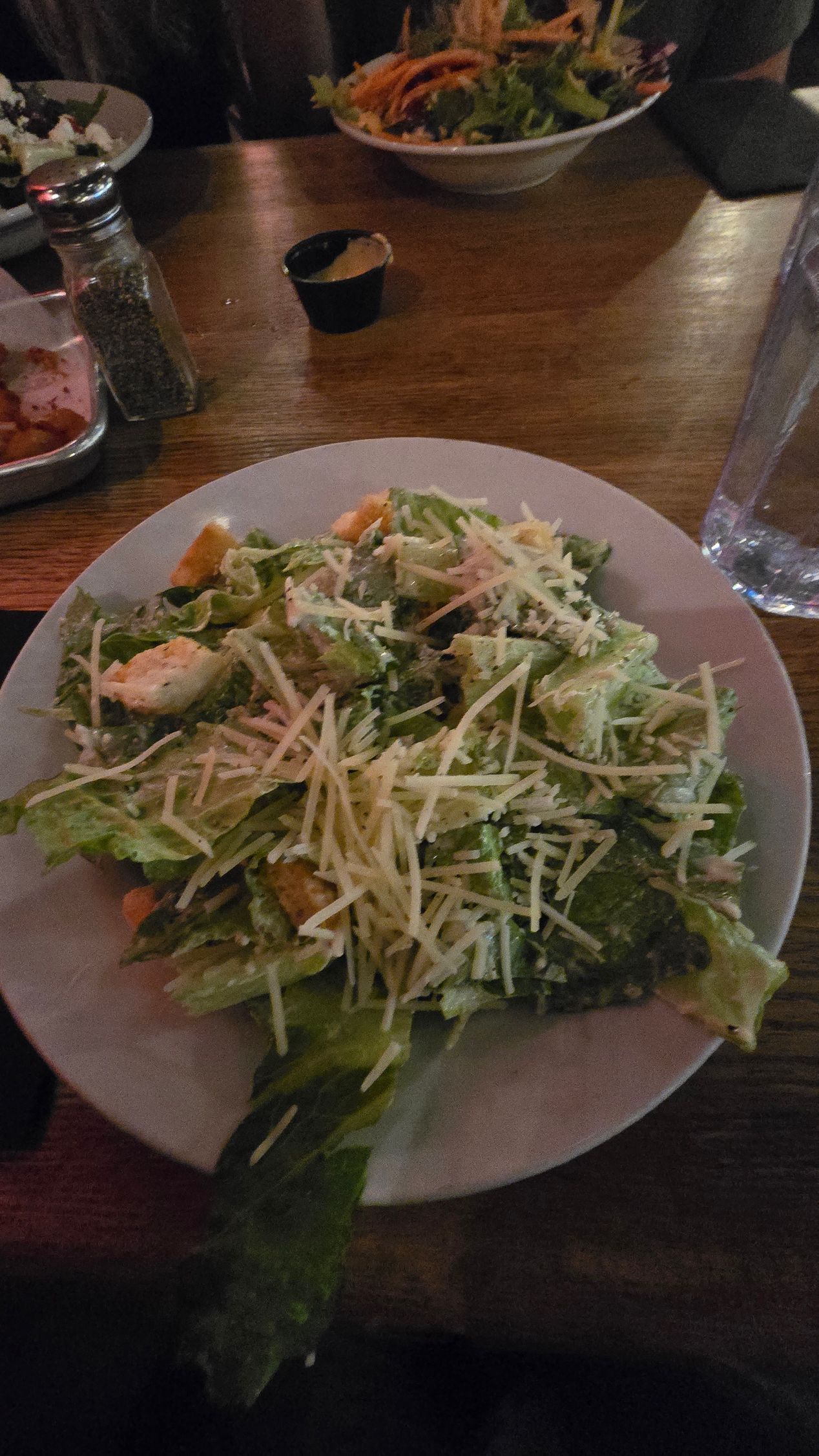 Plate of green salad topped with shredded cheese on a wooden table in a dim restaurant.
