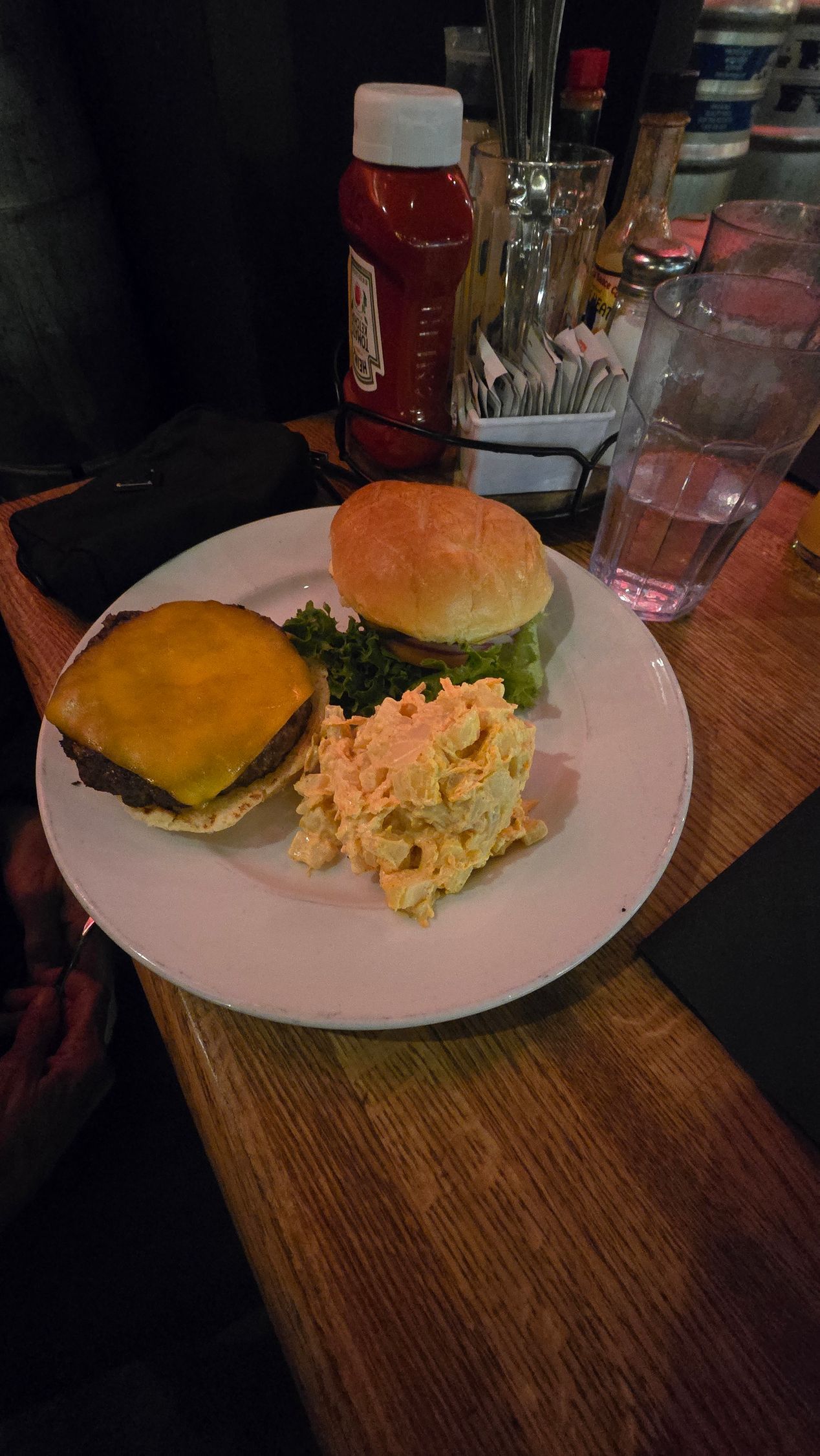Plate with cheeseburger, scrambled eggs, and cheese on a wooden table beside a ketchup bottle and glass.