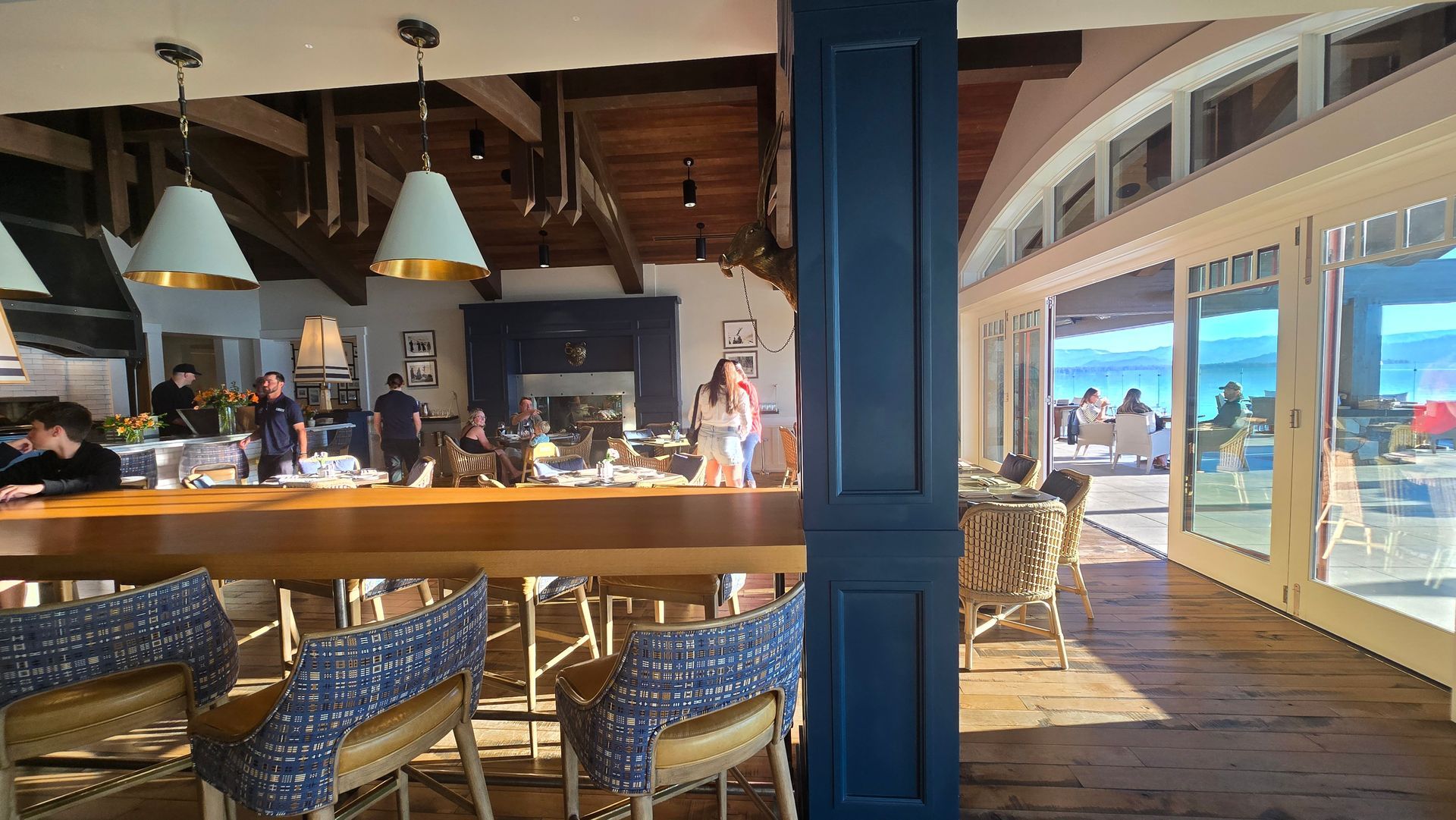 Cozy waterfront restaurant with blue chairs, warm pendant lights, and large windows overlooking the sea at sunset