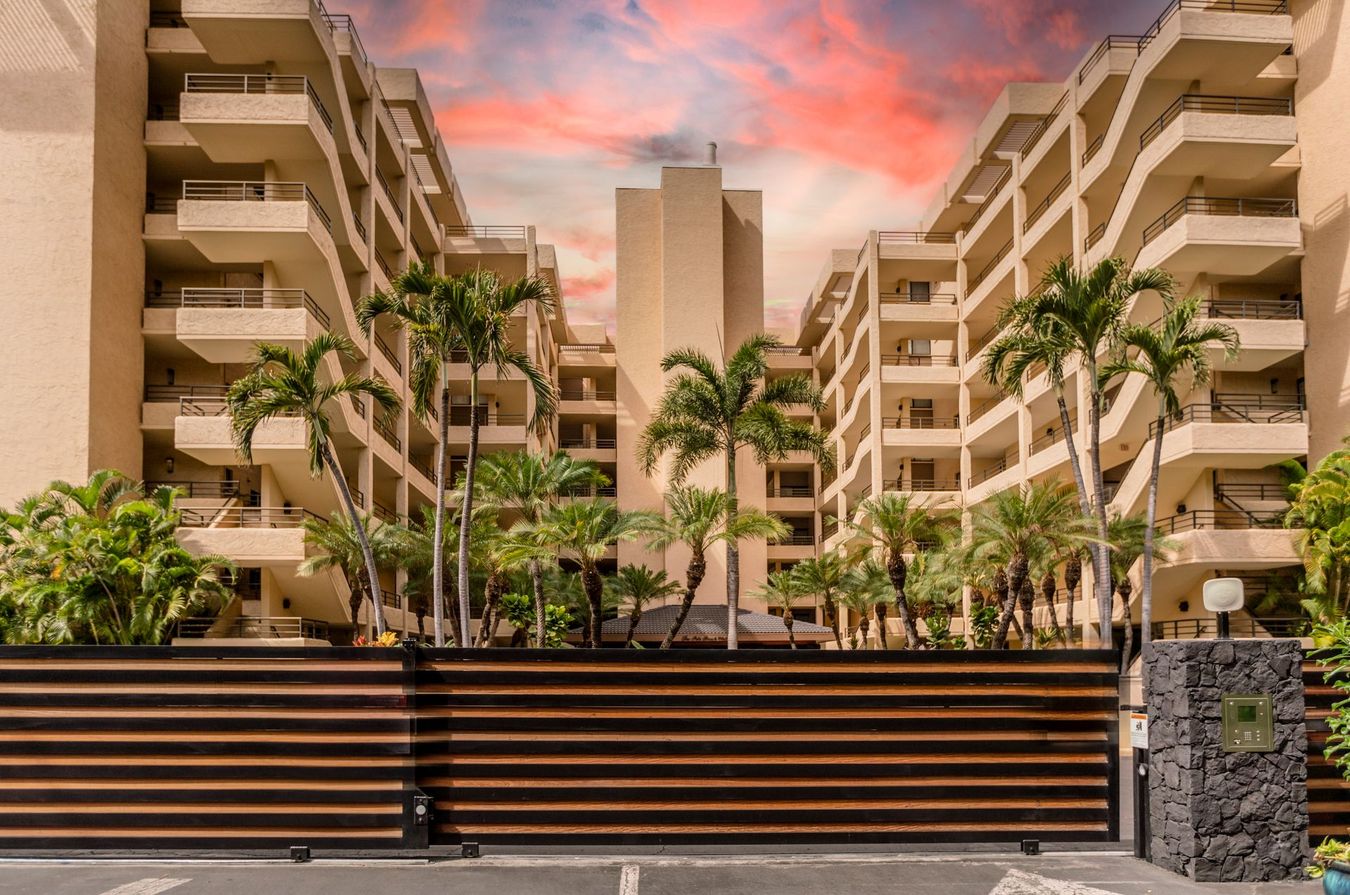 Brown gated entrance to beige apartment buildings with palm trees under a colorful sky.
