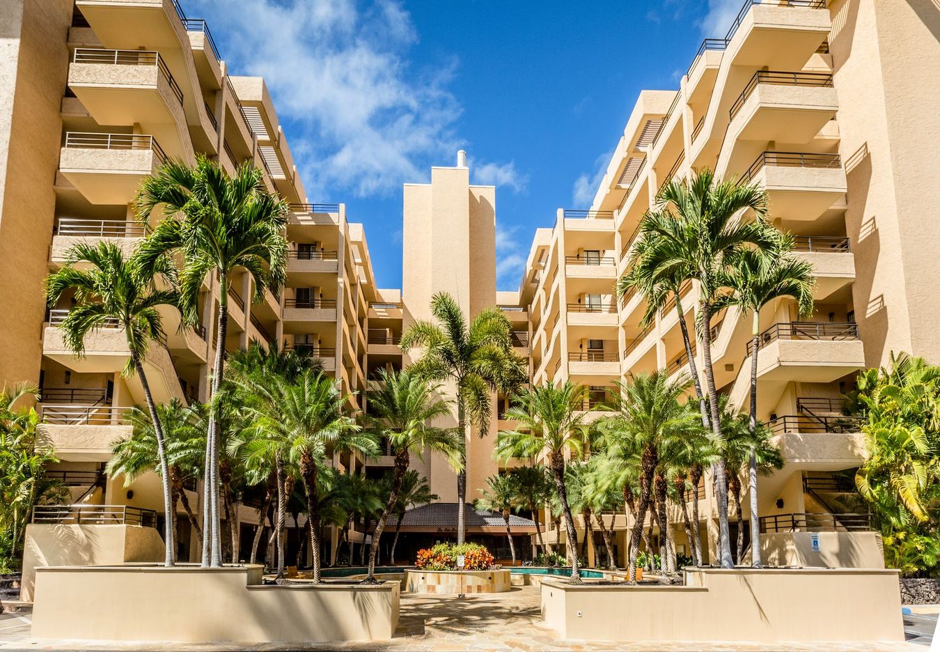 Tan multi-story condo building with palm trees under a blue sky.
