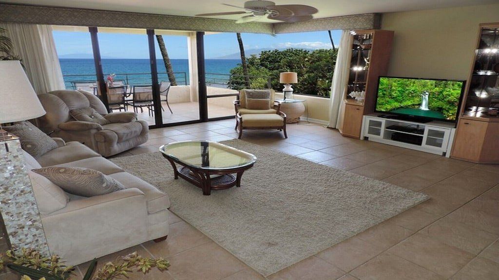 Living room with ocean view: cream-colored couch, armchair, rug, TV, sliding glass doors to patio, and blue water.