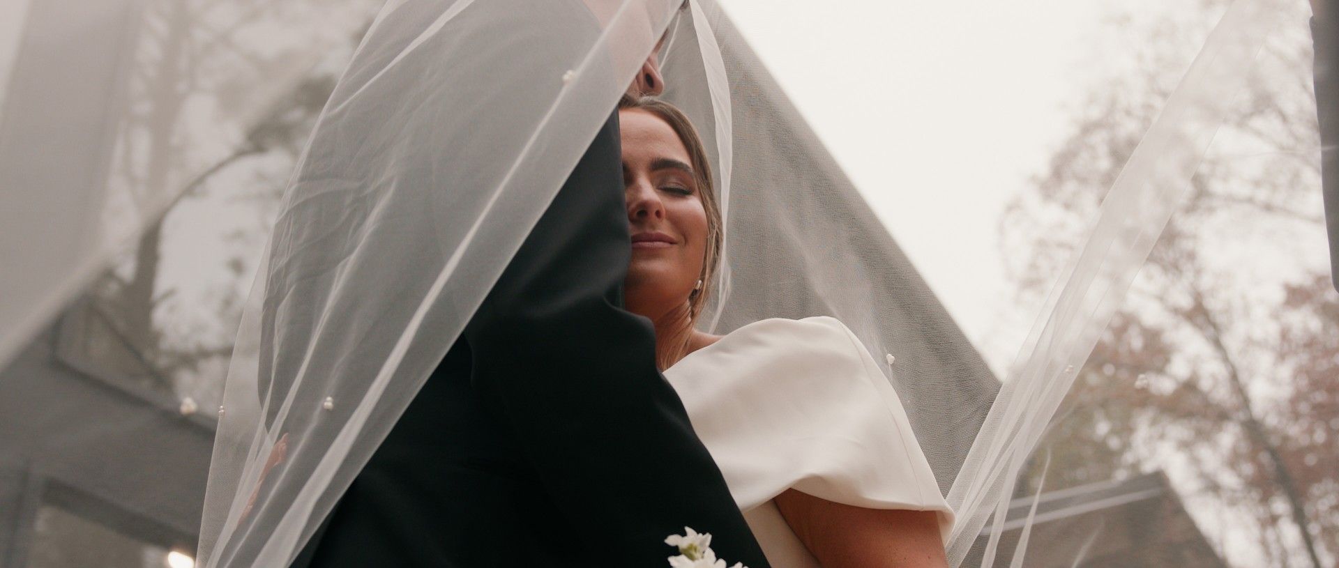 Bride and groom embracing under a veil on their wedding day; outdoors.