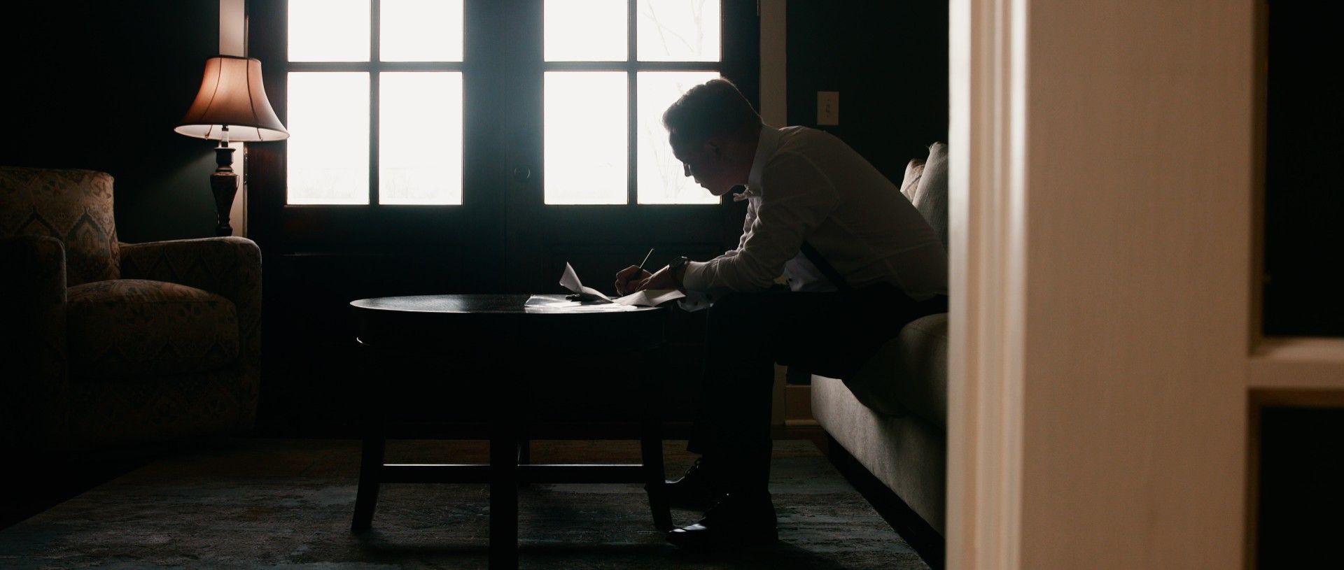 A person sits on a couch, writing at a table in a dimly lit room with a window and lamp.