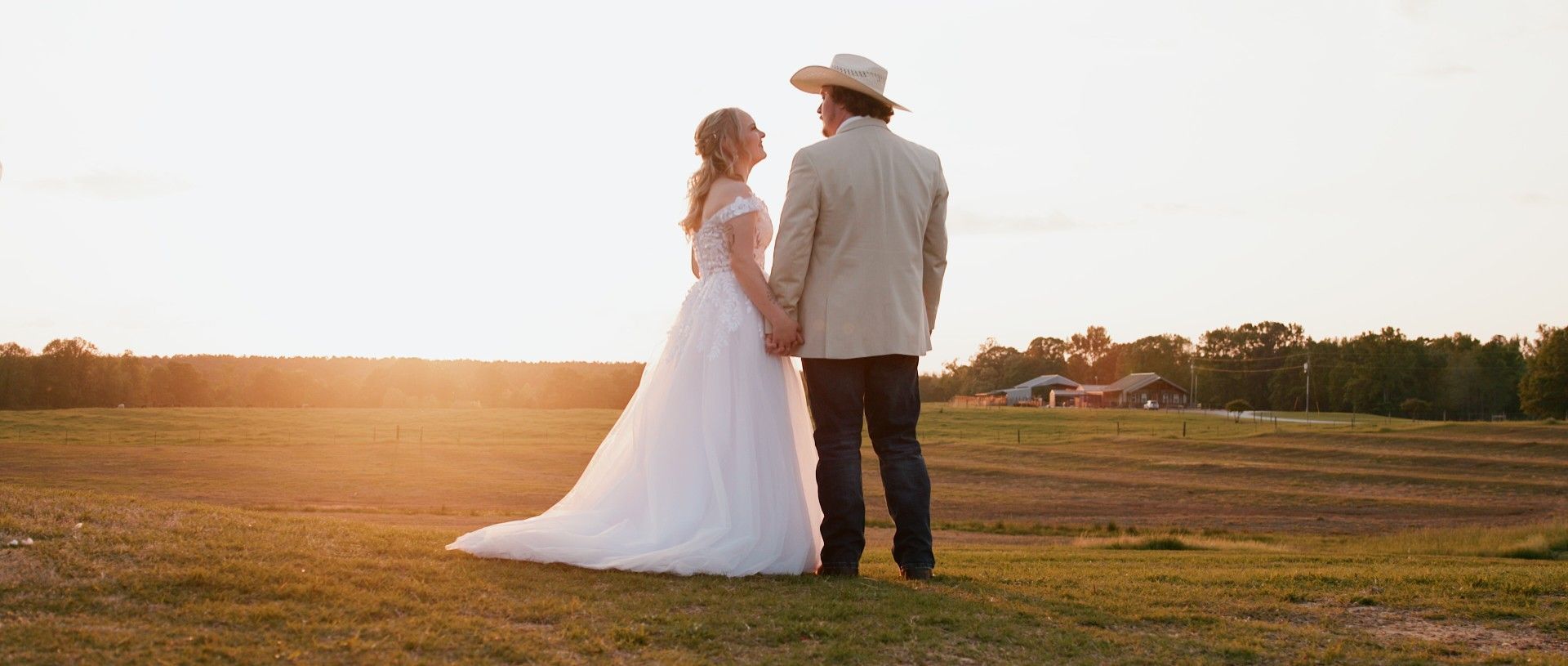 Couple holding hands, standing in a field at sunset, man wearing a cowboy hat, bride wearing a wedding dress.