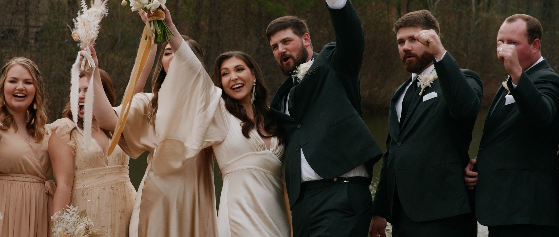 Wedding party celebrating outdoors. Bride and groom raise arms, bridesmaids hold bouquets. Groomsmen fist pump.