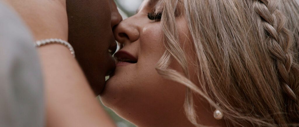 Close-up of two people kissing. The person on the right is blonde with a braid, wearing a pearl earring.