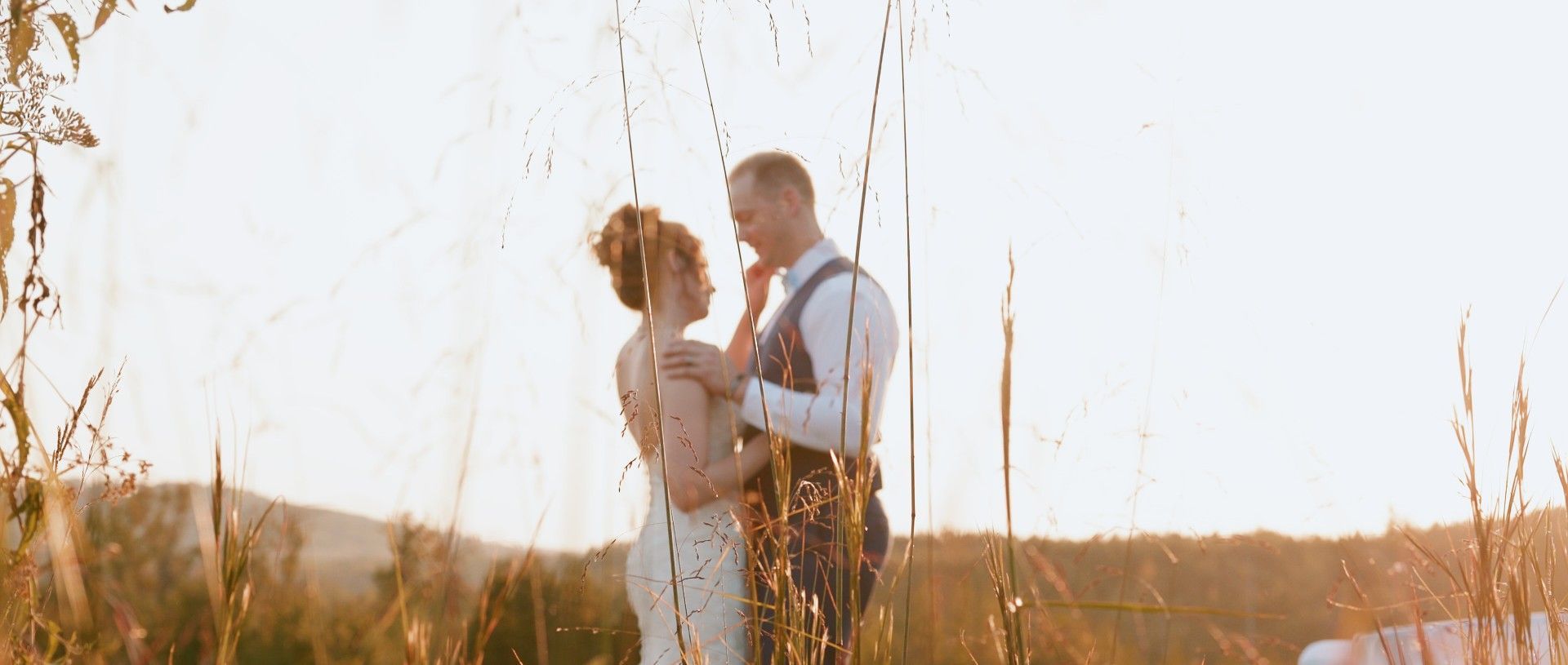 A couple embraces in a sunlit field, wedding attire, warm tones, and golden hour lighting.
