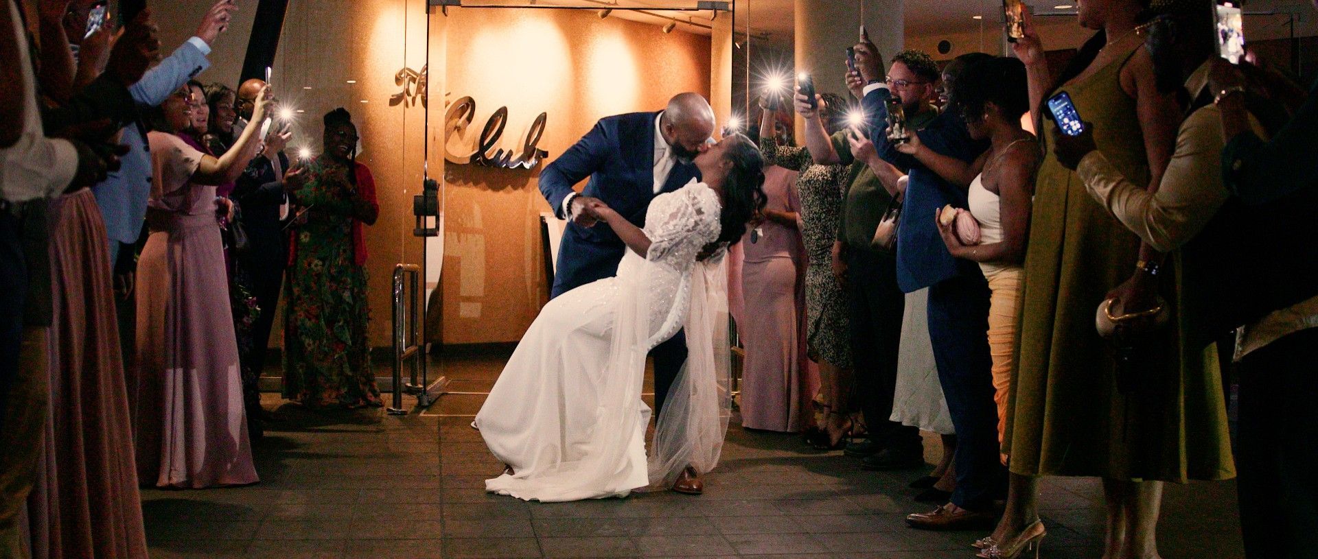 Newlyweds kiss amidst a crowd of guests holding up phones. They exit a building with 