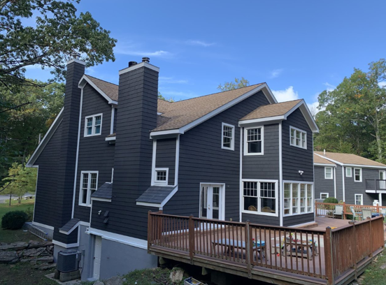 Dark gray two-story house with white trim, wooden deck, and chimney under a blue sky