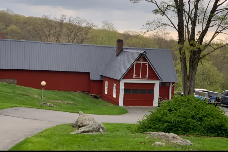 Red barn with gray roof on a grassy hillside, surrounded by trees and a driveway.