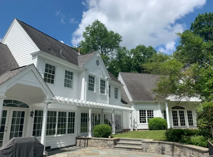 White house with dormers and large porch, set among trees under a bright blue sky