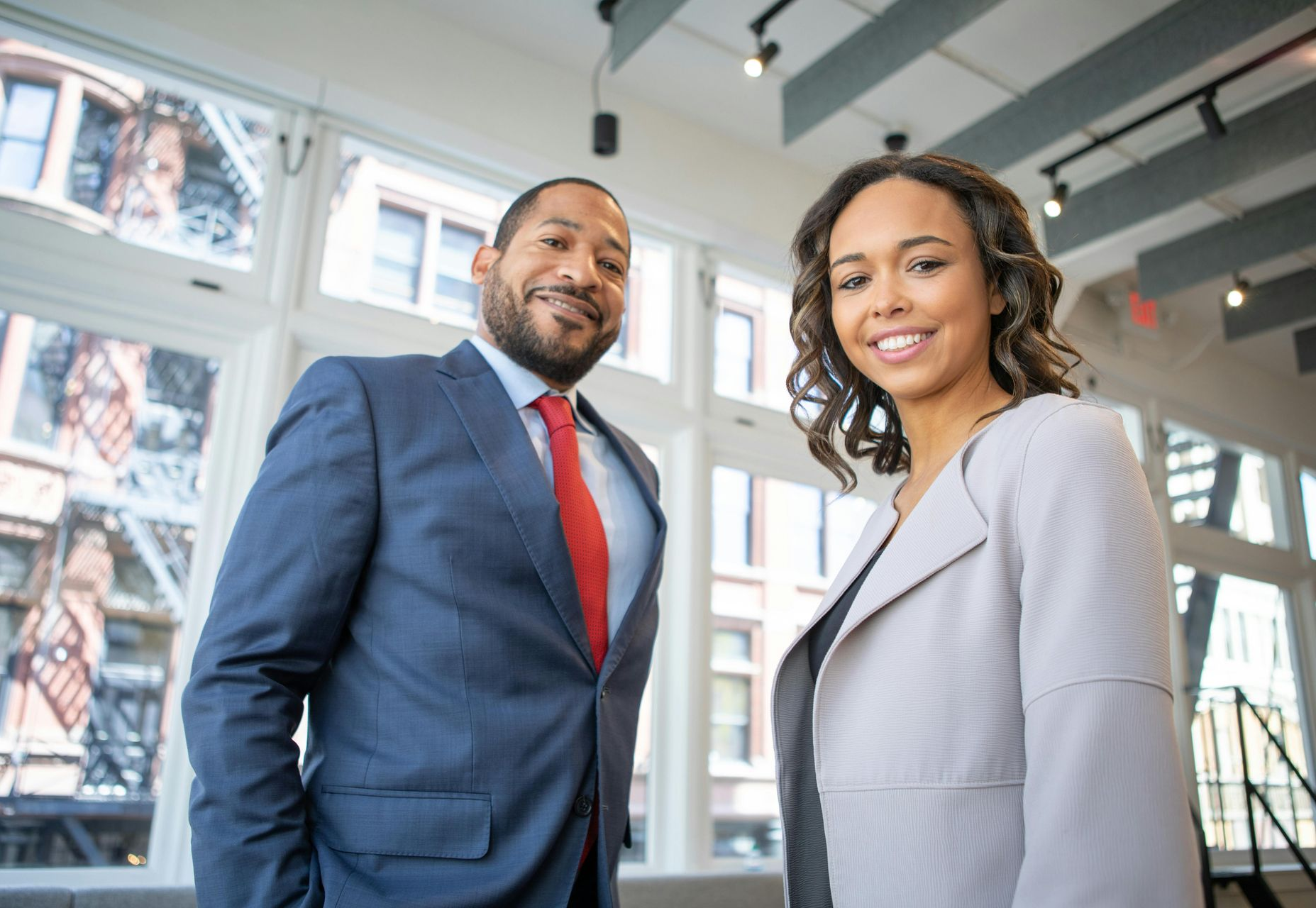 Man in a blue suit with a red tie and a woman in a light gray jacket smiling in a modern office.