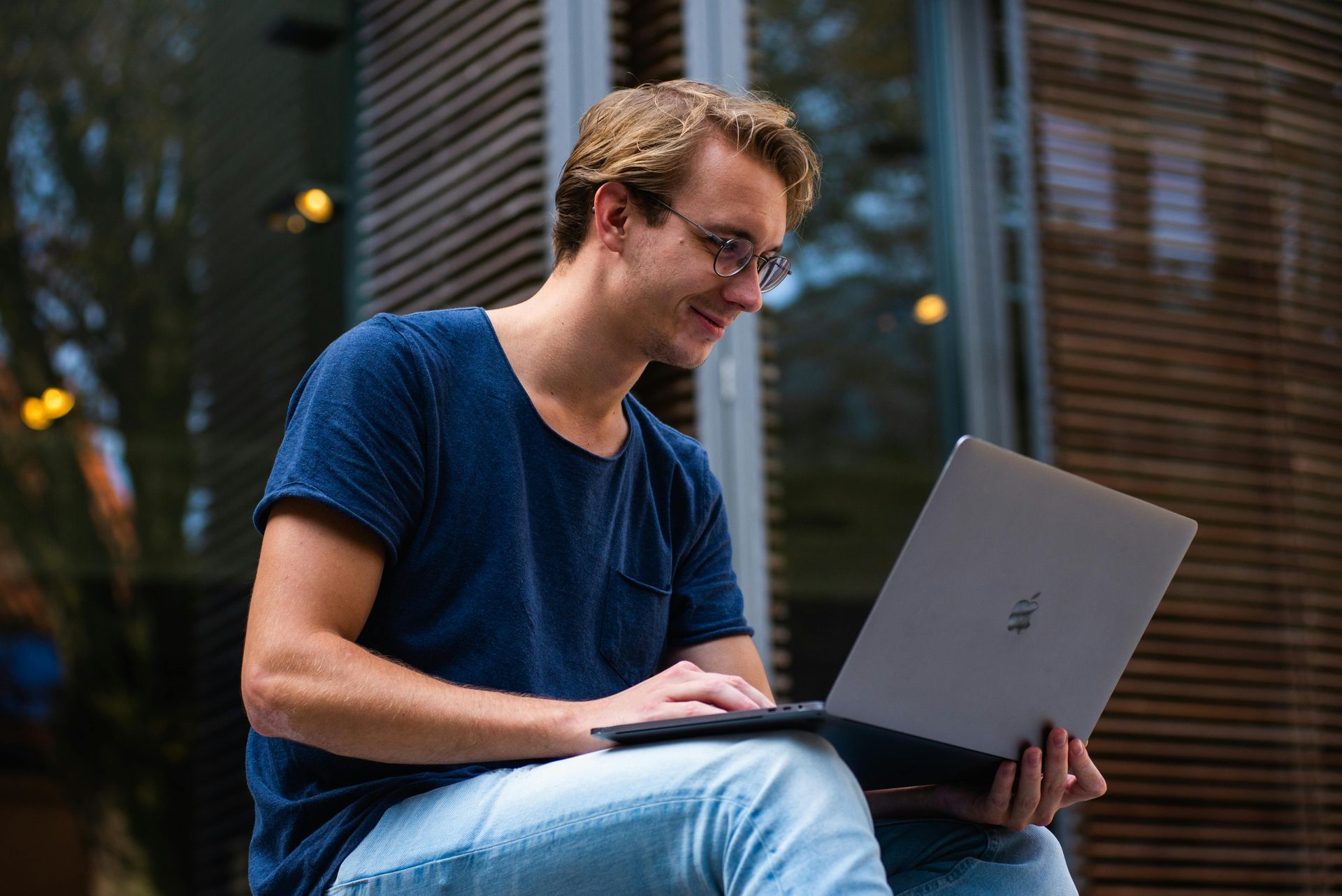 Man wearing glasses and a blue shirt, smiling while using a laptop outdoors.