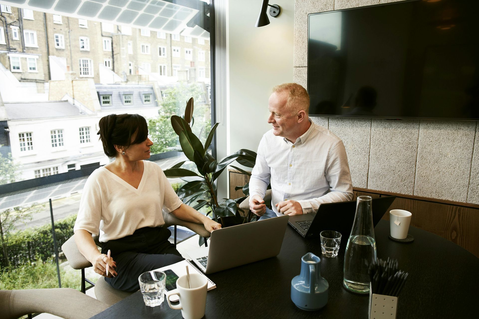 Two people sit at a table in a modern office, working on laptops. A large window offers a city view.