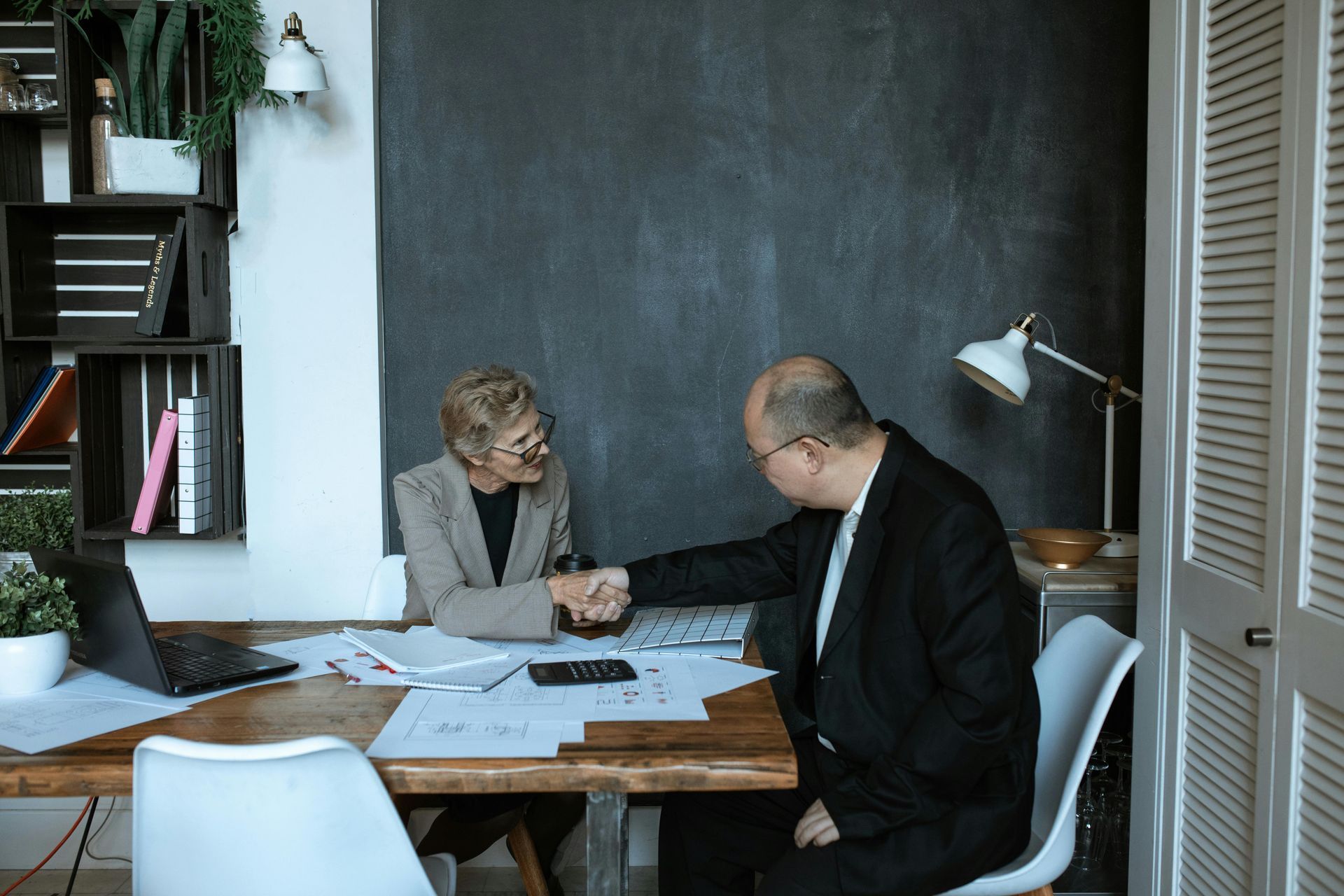 Man and woman in suits shake hands over a desk with papers in an office setting.
