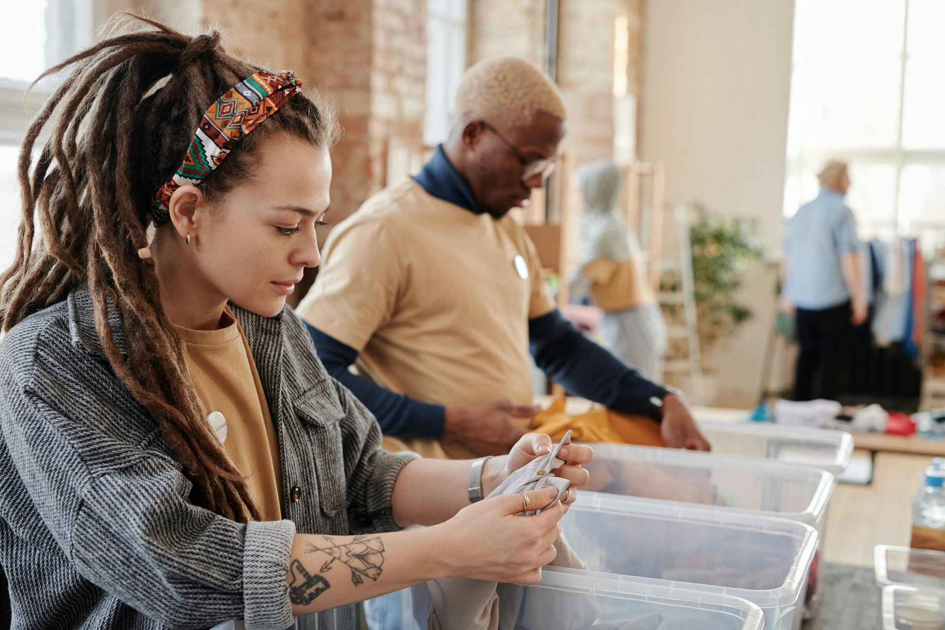 People sorting items into bins in a brightly lit room. Woman with dreadlocks examines items, others sort in background.
