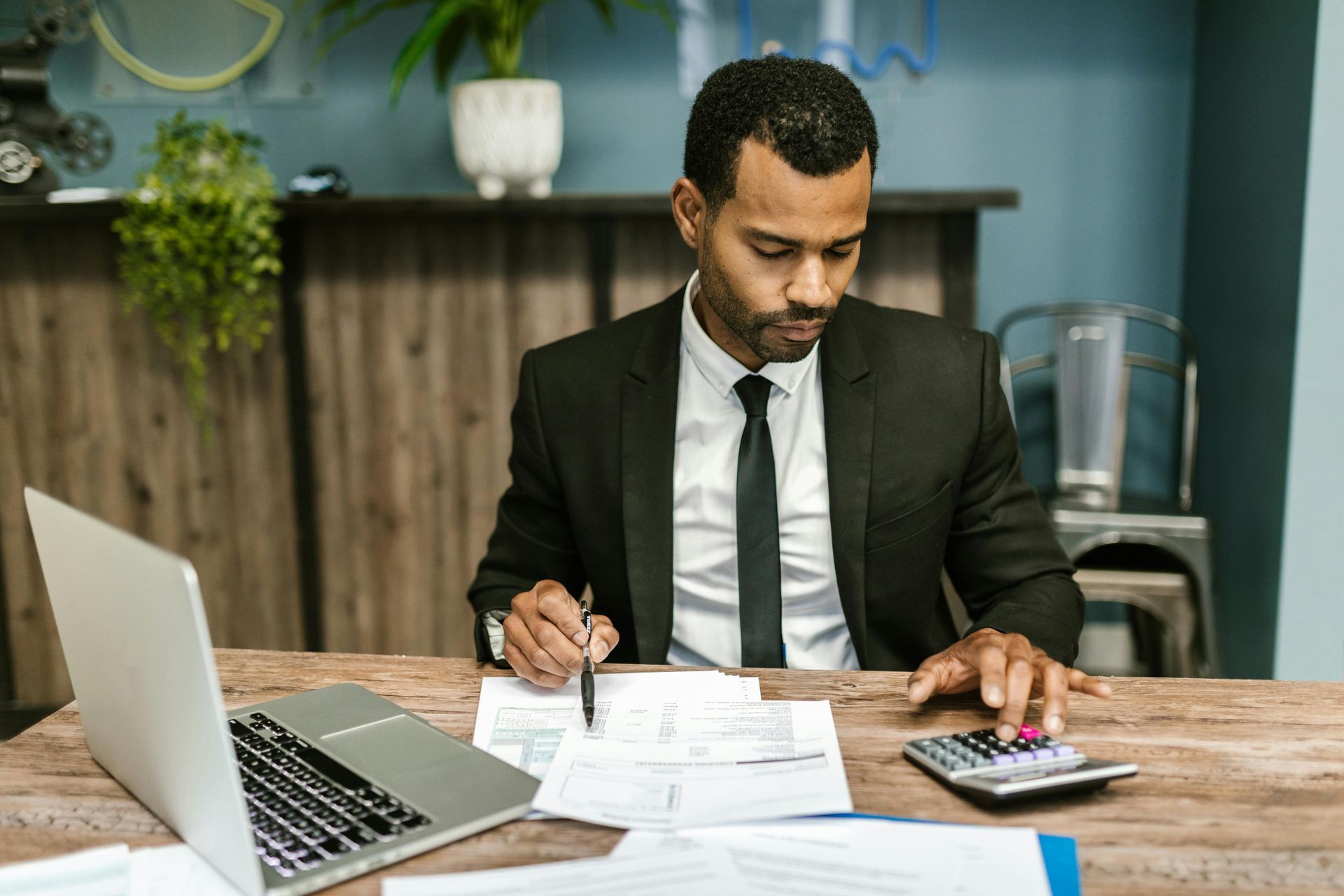 Man in suit using calculator and laptop, reviewing documents at a wooden desk in office.