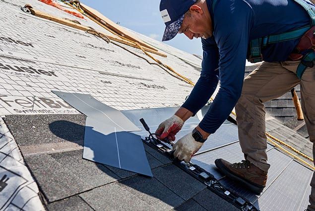 Worker installing shingles on a roof with a power tool and gray flashing sheets