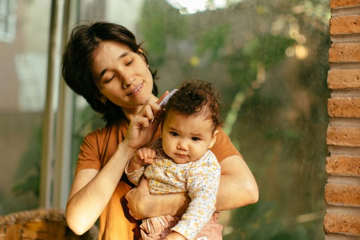 A woman is holding a baby in her arms and brushing her hair.