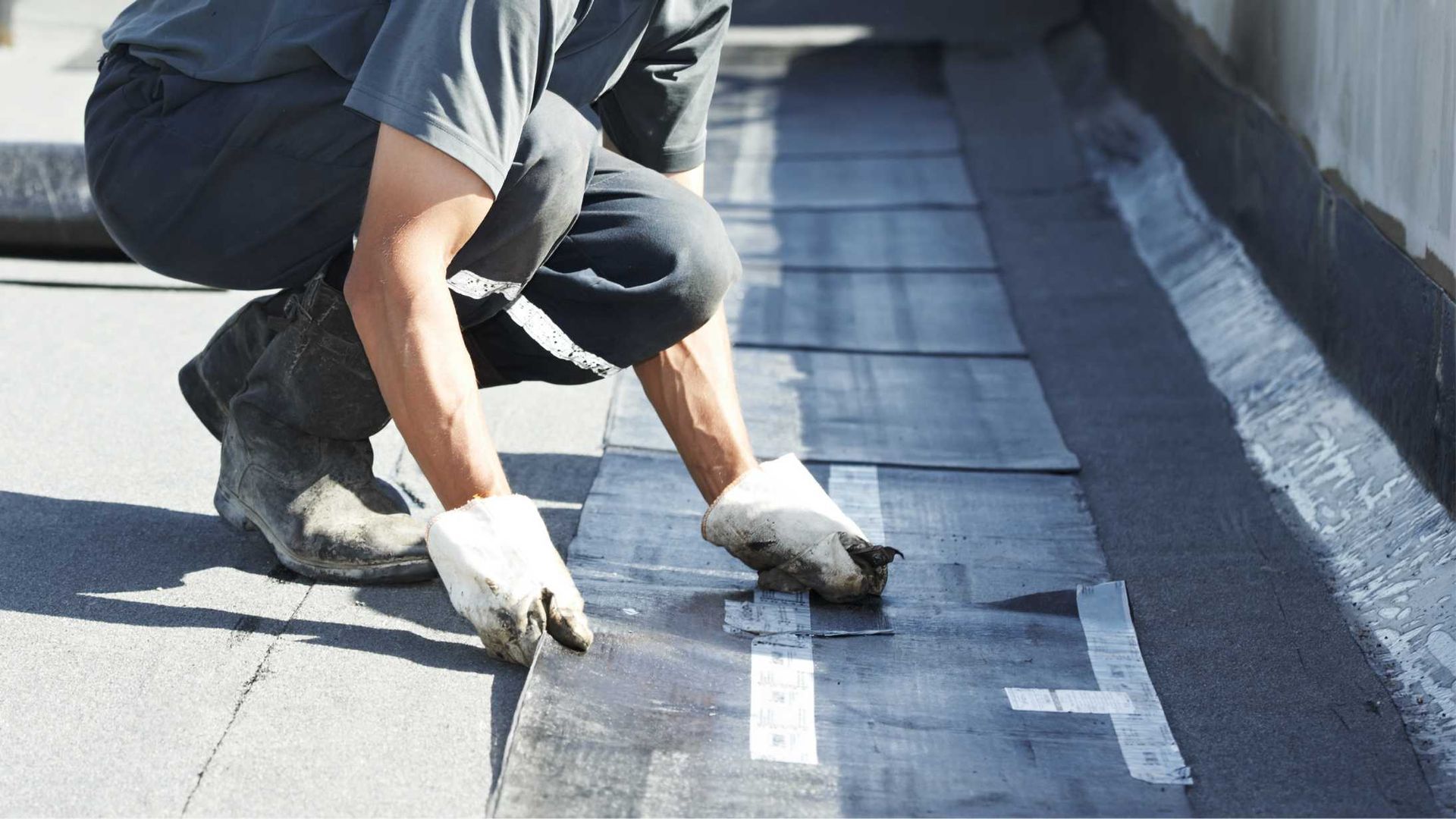 A worker in work clothes and gloves installs black roofing material on a flat roof.