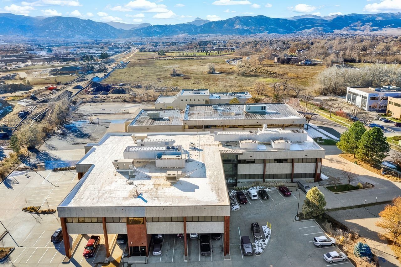 An aerial view of a multi-story office building with a flat roof, parking lot, and distant mountain range.