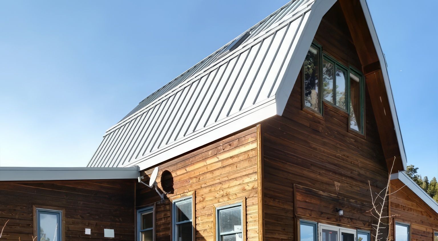 A wooden house with a steep, light-colored metal roof under a bright blue sky.