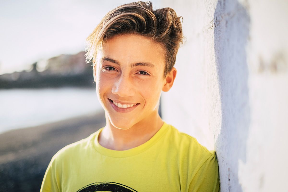 A teenaged boy in a yellow shirt is smiling, standing by the ocean.