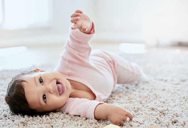 Baby lying on a light-colored carpet, smiling, with one arm raised. Wearing a pink outfit.