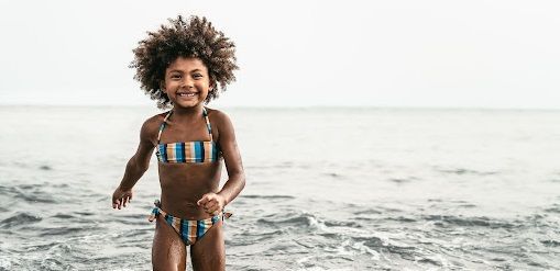 Young girl in a bathing suit walking in the ocean, smiling at the camera.