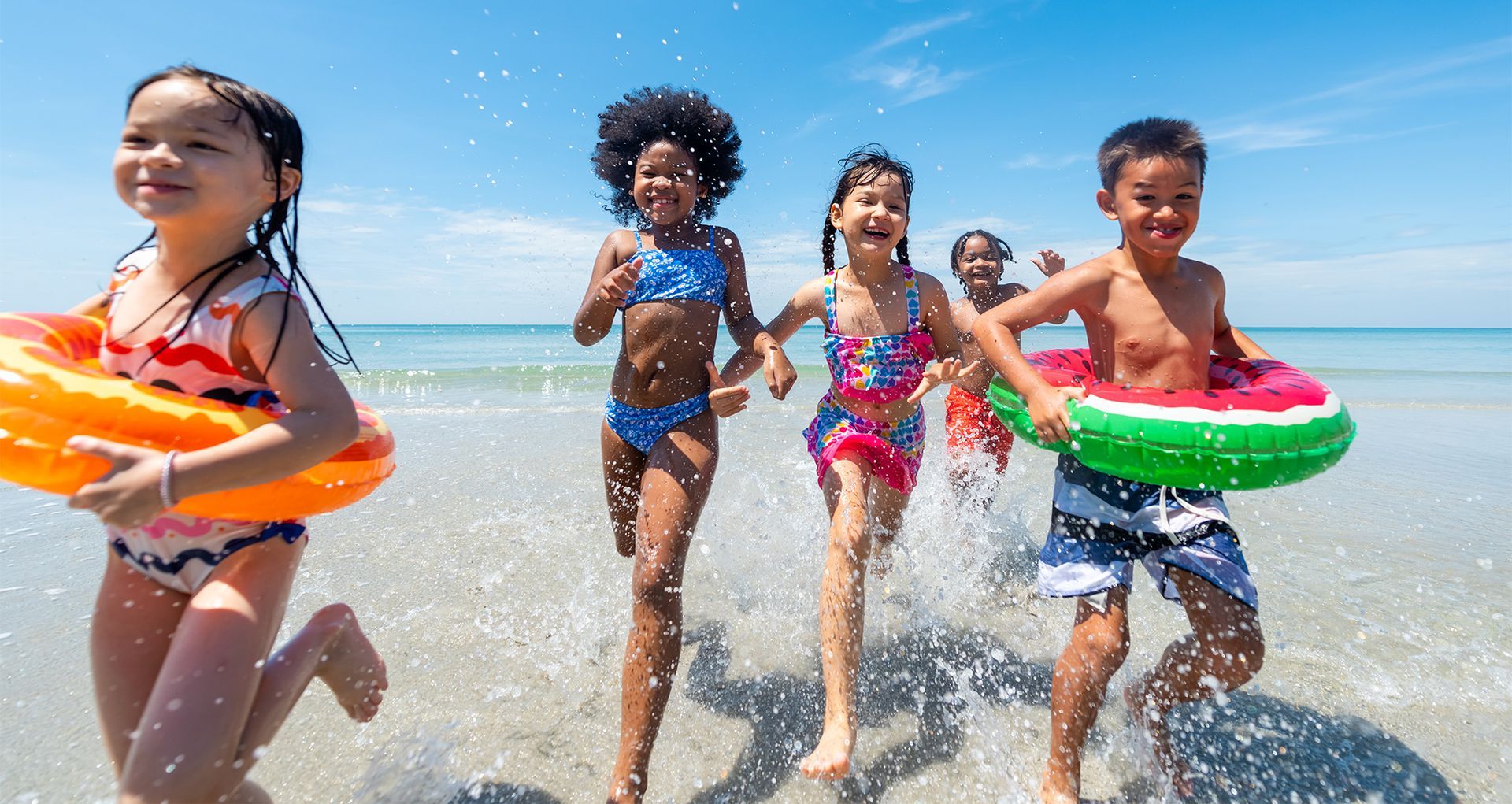 Five children in suits and floaties are running on the beach. They are smiling.