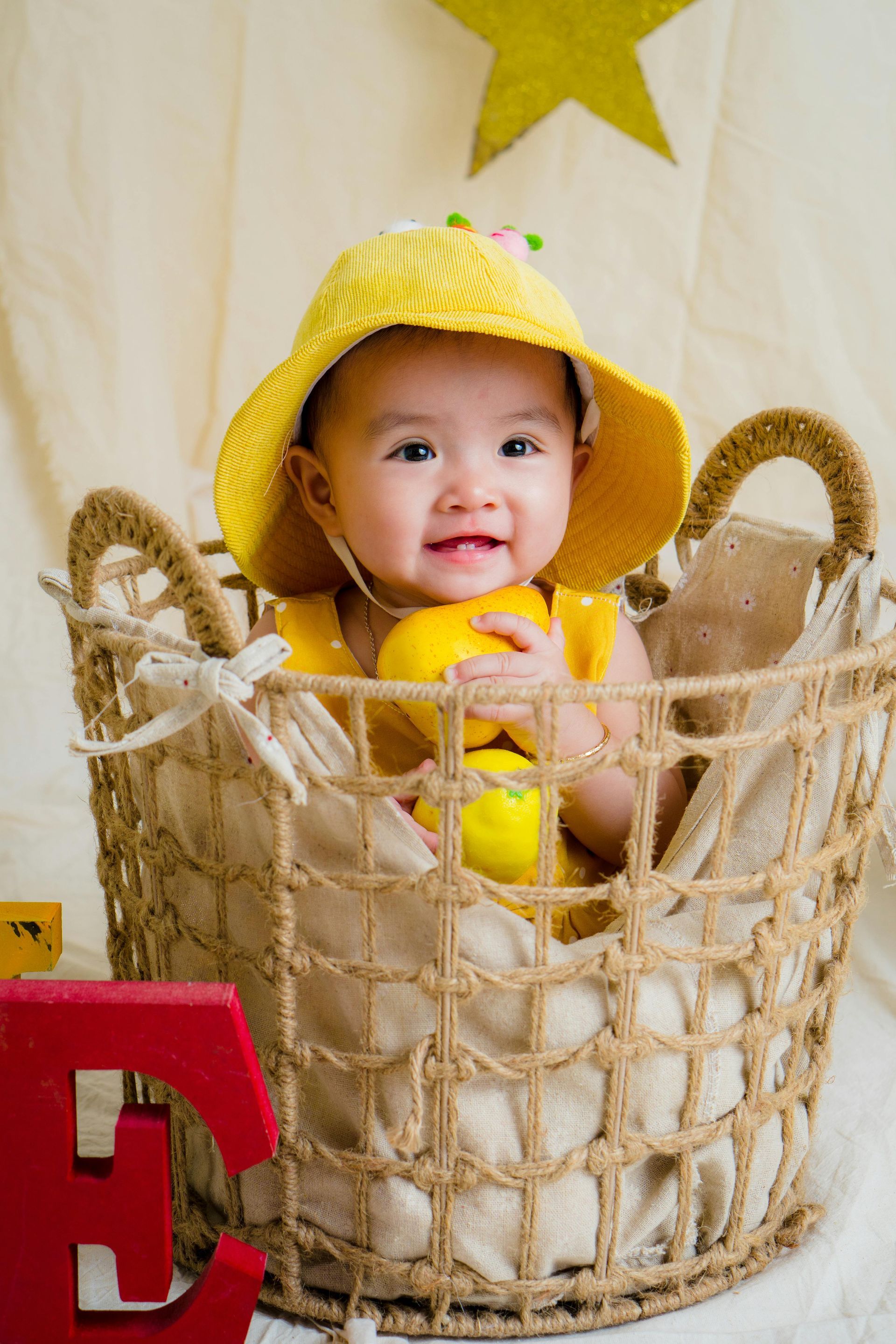 Baby in yellow hat and dress smiles, holding lemons in a wicker basket.
