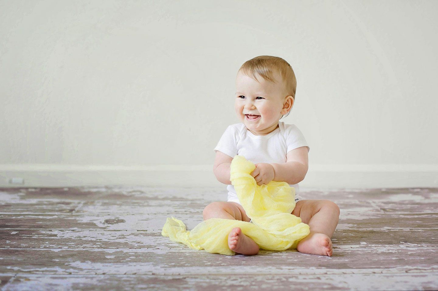 Smiling baby in white onesie sits on a wood floor, holding a yellow cloth.