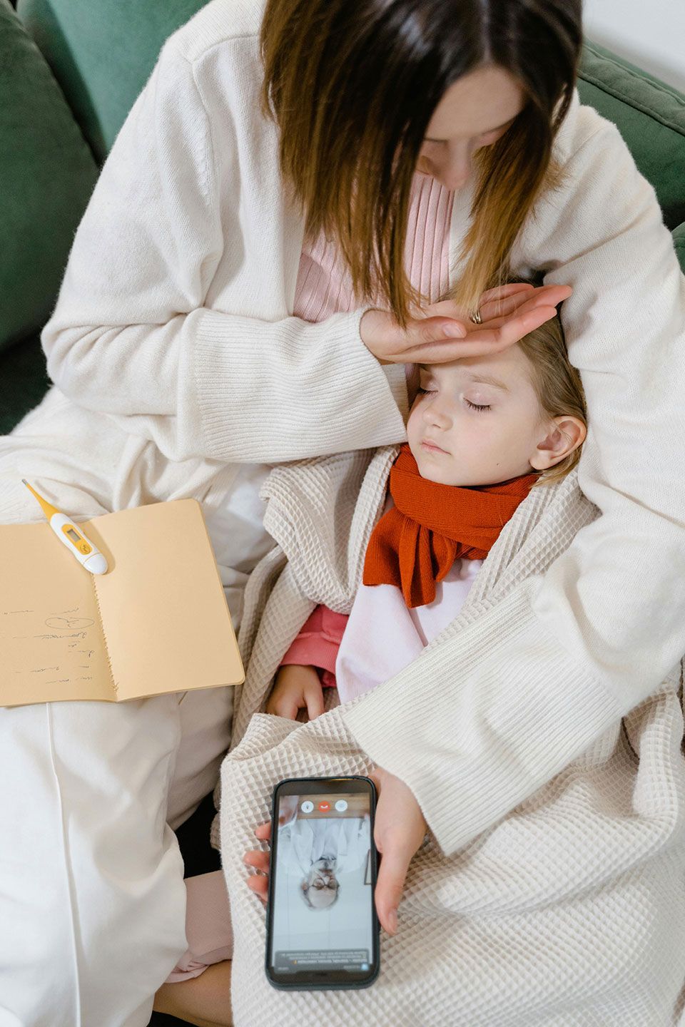 Woman checking a child's forehead. The child is wrapped in a blanket, wearing a scarf. Phone and notepad are present.