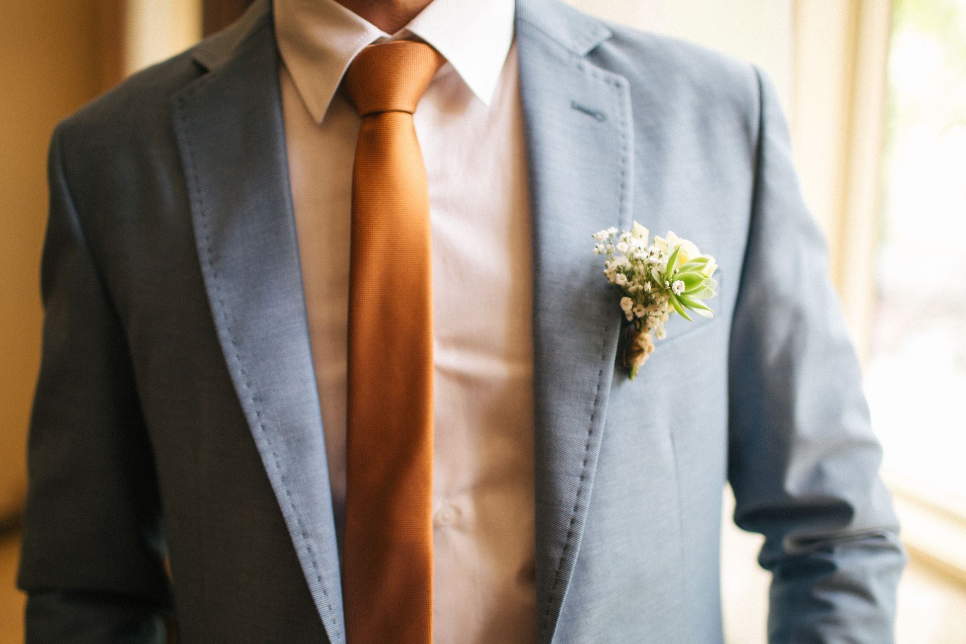 Man in light blue suit, orange tie, and white shirt with a floral boutonniere.