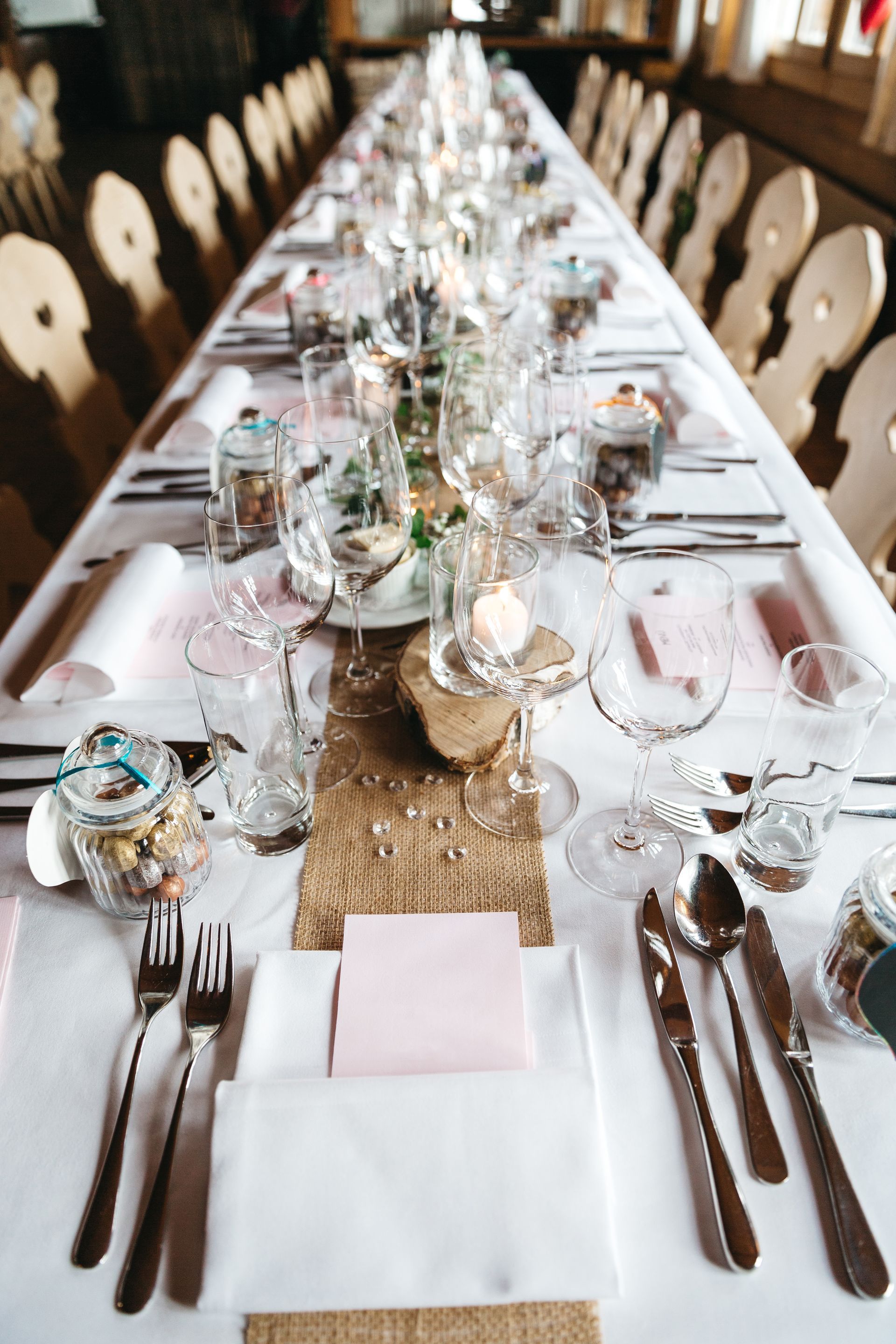 Long wooden tables set for a wedding reception in a forest; floral centerpieces, candles, and blue napkins.