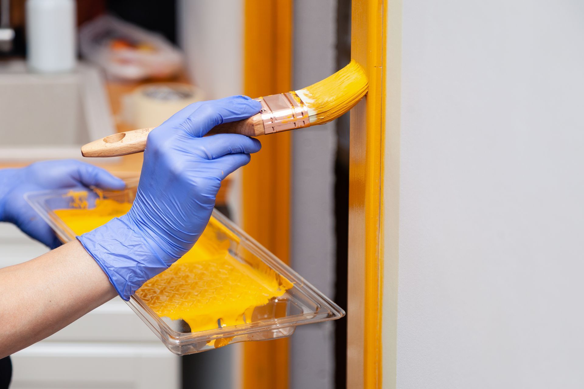 Person in blue gloves painting a yellow door frame with a brush and paint tray.