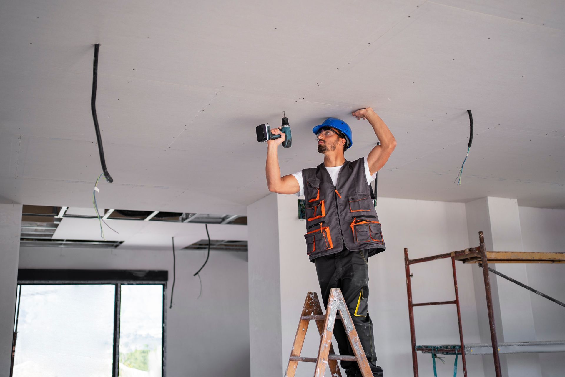 A man is standing on a ladder using a drill to fix a ceiling.
