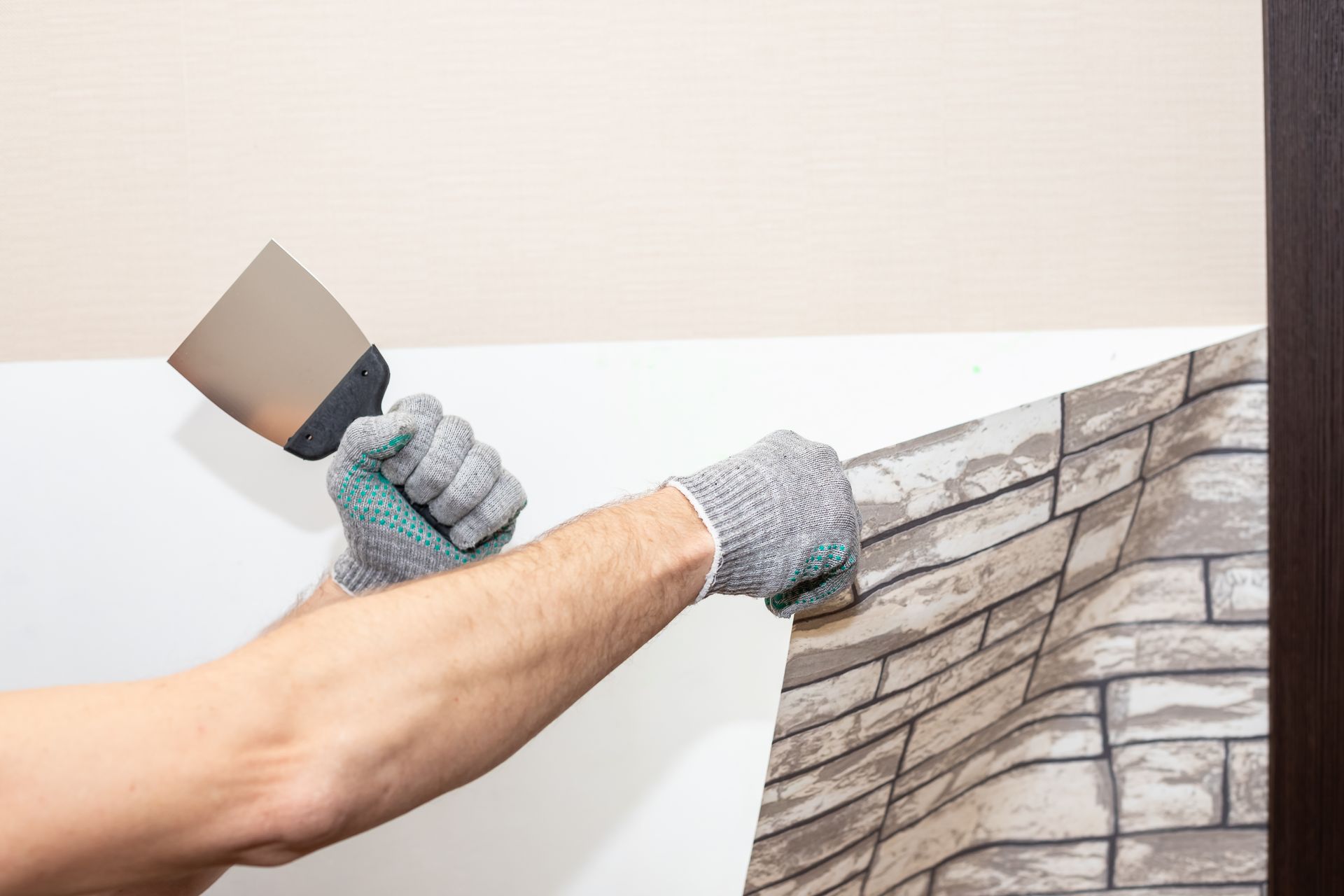 A man is using a spatula to remove a brick wallpaper from a wall.