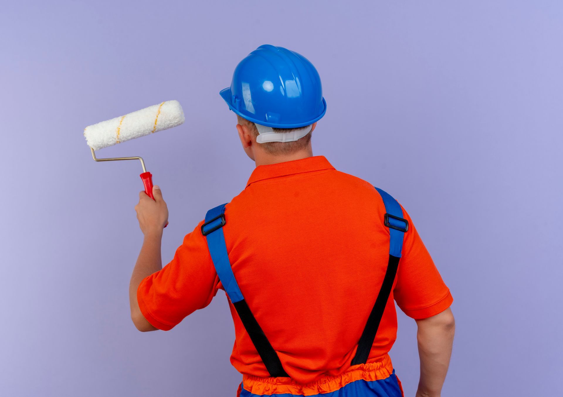 A man in an orange shirt and blue overalls is holding a paint roller.