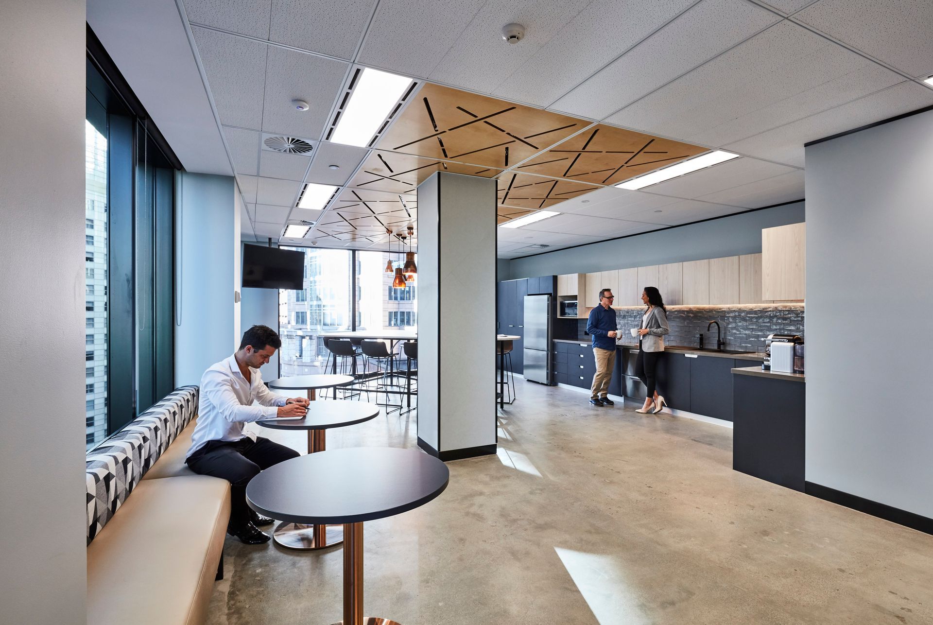 Office space with a seated person working at a table, people in kitchen area, window views.