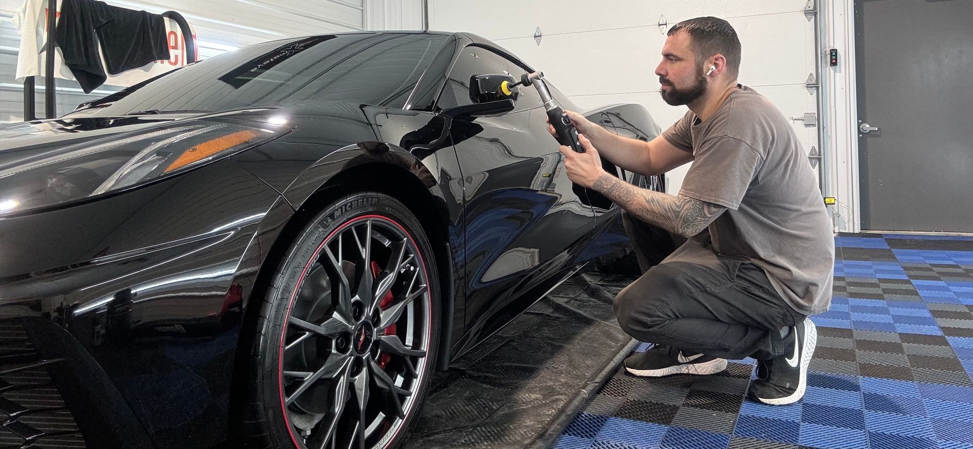 A person cleans a black sports car in a garage with a blue and gray tiled floor.