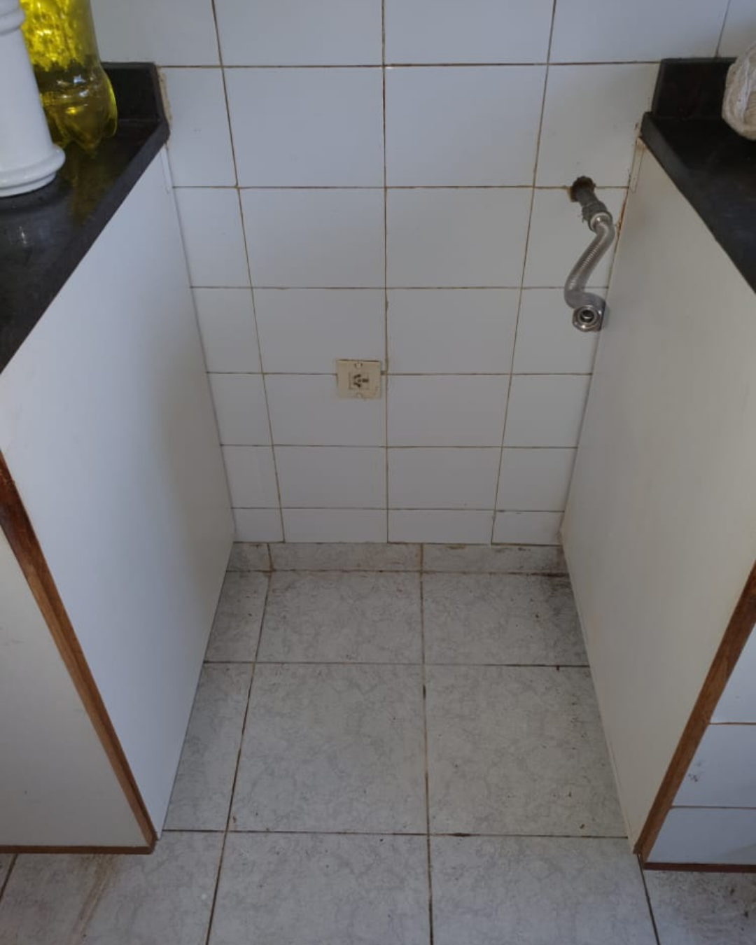 Pequeña área de cocina con gabinetes blancos y paredes y piso de azulejos; grifo en la pared.