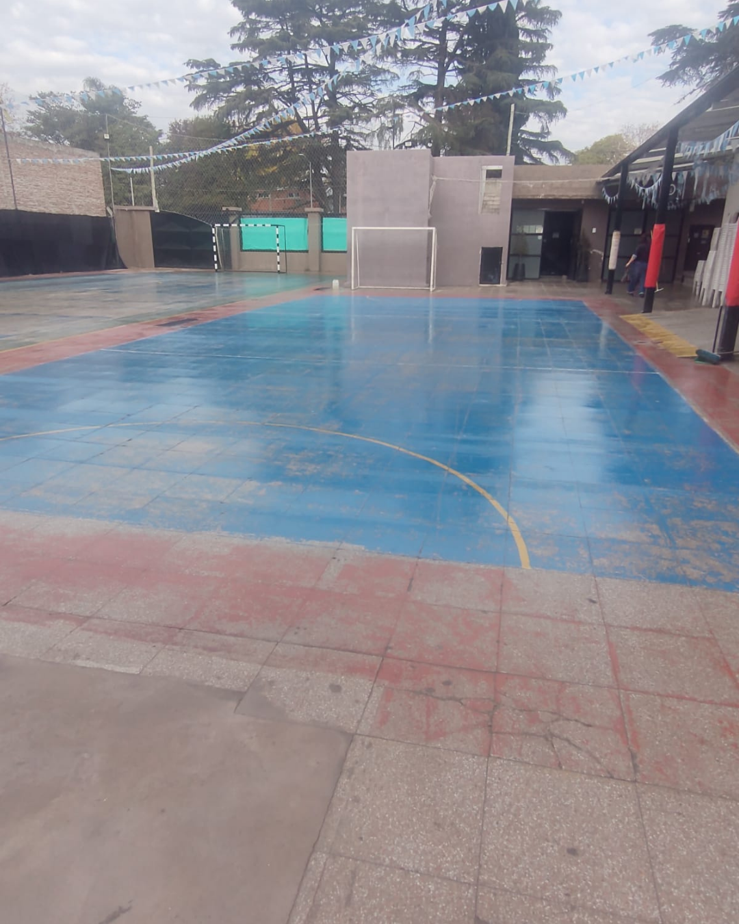 Cancha deportiva pavimentada en azul y rojo con portería de fútbol. Árboles y edificios al fondo.