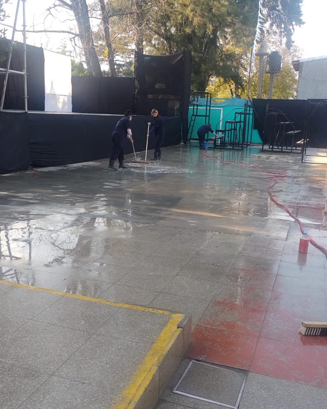 Trabajadores lavando un área exterior de concreto húmeda con mangueras y escobas; escenario al fondo.