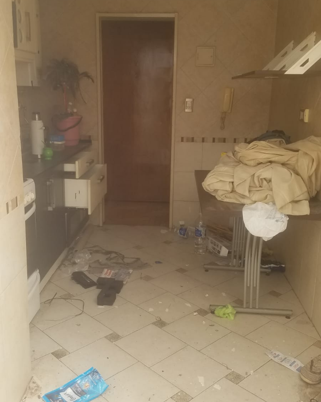 Cocina desordenada con escombros en el suelo, mesa cubierta de ropa y puerta abierta.
