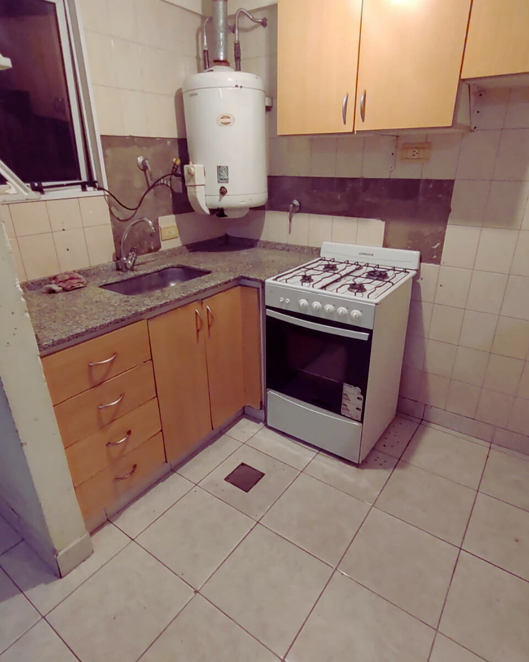 Pequeña cocina con estufa, fregadero, calentador de agua y gabinetes de madera clara.