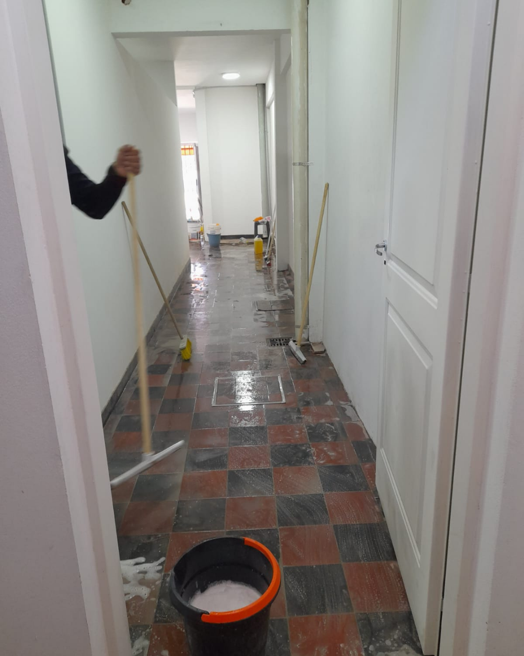 Persona fregando un pasillo con suelo de baldosas a cuadros. En primer plano, un cubo con agua jabonosa.