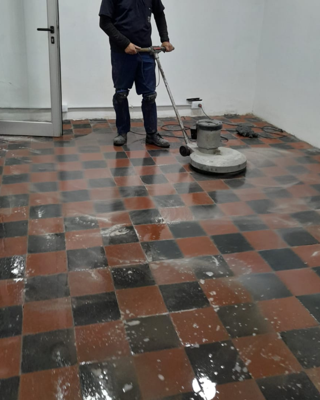 Una persona usa una pulidora de pisos sobre un suelo de baldosas cuadriculadas. El suelo está mojado.